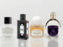 A banner-style photograph featuring four unique perfume bottles arranged on a glossy white reflective table. From left to right: Fulton & Roark, Xyrena, KST Scent, and Krigler.