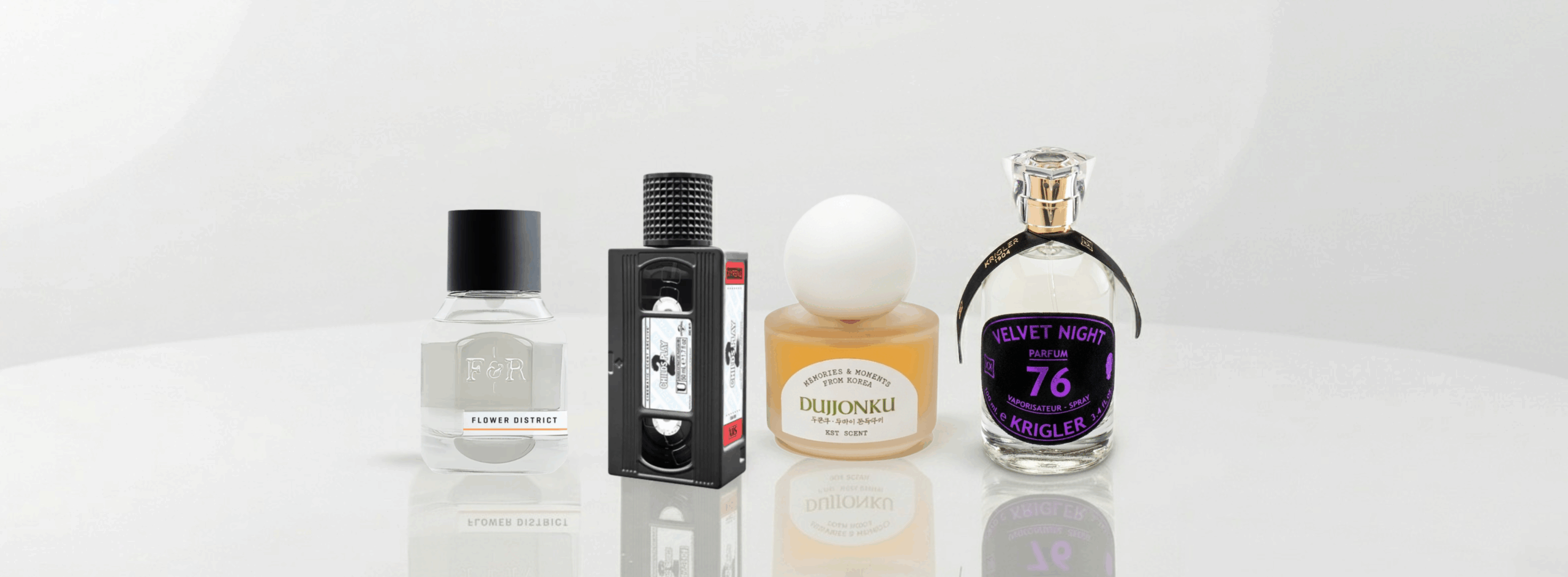 A banner-style photograph featuring four unique perfume bottles arranged on a glossy white reflective table. From left to right: Fulton & Roark, Xyrena, KST Scent, and Krigler.